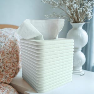 May include: A white tissue box with a ridged design, holding a stack of white tissues. The box is square and sits on a white surface, next to a white vase with dried flowers.