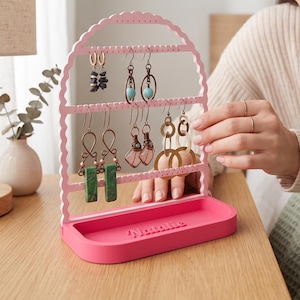 May include: A light pink arched earring stand with three horizontal bars featuring small holes for hanging jewelry. The base is a solid dark pink rectangular tray with the name Natalie embossed on the front. Several pairs of earrings are displayed.