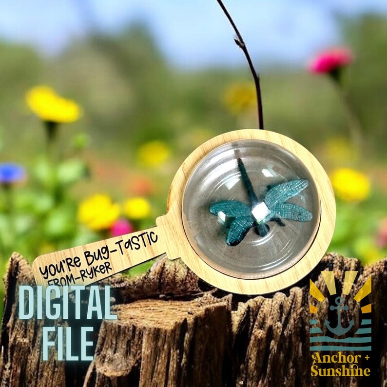 May include: A wooden magnifying glass with a clear plastic lens, showing a blue dragonfly inside. The handle is engraved with the text "You're Bug-Tastic from-Ryker". The magnifying glass is sitting on a tree stump with a blurred background of flowers.