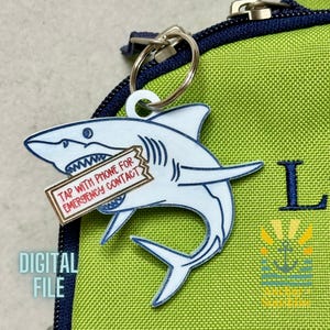 Op de afbeelding: Een wit en blauw haai-vormige sleutelhanger met de tekst "TAP WITH PHONE FOR EMERGENCY CONTACT" op een klein label. De sleutelhanger is bevestigd aan een groene tas met een rits en de letter "L". De woorden "DIGITAL FILE" zijn ook zichtbaar.