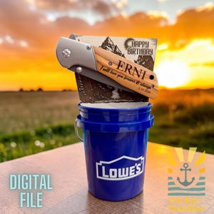 Può includere: Un coltello tascabile in legno personalizzato con una lama argentata, inciso con il nome "ERNI" e un messaggio, esposto con un biglietto di "Buon Compleanno". Il coltello poggia su un secchio blu Lowe's. Le parole "DIGITAL FILE" sono visibili.