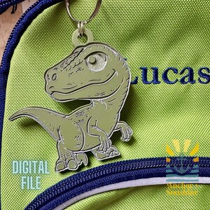 May include: A green dinosaur keychain with a smiling face, attached to a green backpack. The backpack has a blue zipper and the name "LUCAS" embroidered in blue. The words "DIGITAL FILE" are also visible.