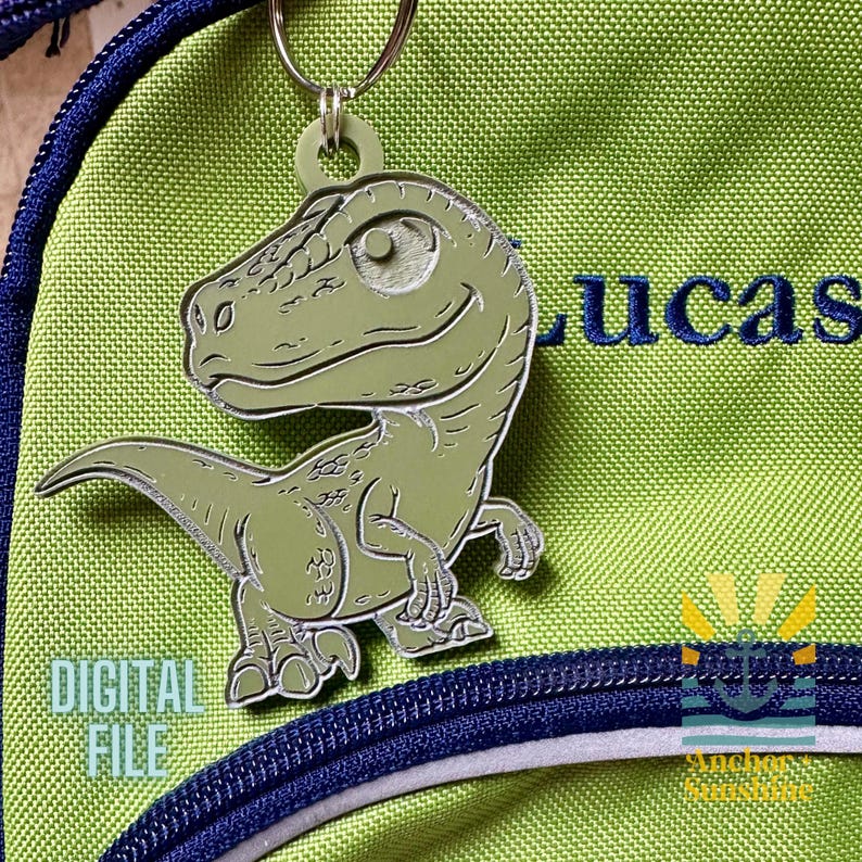May include: A green dinosaur keychain with a smiling face, attached to a green backpack. The backpack has a blue zipper and the name "LUCAS" embroidered in blue. The words "DIGITAL FILE" are also visible.