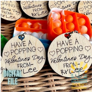 May include: Wooden Valentine's Day ornaments with the text "Have a Popping Valentine's Day From - Rylee". The ornaments have a heart-shaped cutout and are attached to a keychain. The image also shows a pop-it toy.