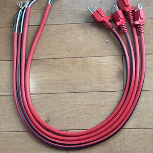 May include: A collection of red electrical extension cords with black stripes, and silver prongs. The cords are arranged in a loop, with the plugs on one end and the wires on the other.
