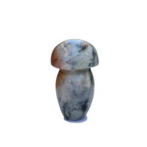 May include: A mushroom-shaped decorative object with a rounded cap and a cylindrical stem. The object is made of a stone material with a marbled pattern of gray, white, and brown hues. The mushroom is standing upright on a white surface.