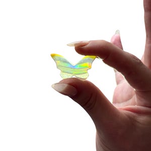 May include: A small, iridescent butterfly-shaped charm with yellow accents. The charm reflects a rainbow of colors and is held between two fingers against a white background. The charm is likely a decorative item or accessory.
