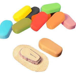 Könnte beinhalten: Eine Vielzahl von bunten, ovalen Objekten, einige mit glatter Oberfläche und andere mit strukturierter Oberfläche. Ein kleiner, rechteckiger, beiger Patch mit einem kleinen, weißen, rechteckigen Gerät mit dem Text "Dexcom G6".