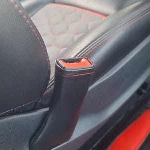 May include: Close-up of a black car seat with red stitching and accents. The seat features a geometric pattern in the center and a black seat belt buckle with a red release button.