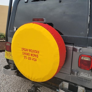 May include: A yellow and red spare tire cover on a gray Jeep. The cover has the text "ORGAN MOUNTAIN CANVAS WORKS 575-208-4124" printed on it. The Jeep has a New Mexico license plate.