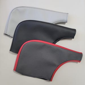 May include: Three automotive accessories in a stack. The top one is light gray, the middle one is black, and the bottom one is dark gray with a red trim. All have a curved shape with a stitched edge.