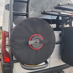 May include: A black spare tire cover with a red accent ring is mounted on the back of a white van. A black ladder is attached to the back of the van. The tire cover has the word "OIL" on it.