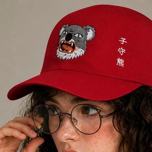 Può includere: Un cappellino da baseball rosso brillante con un ricamo raffigurante il muso di un koala con la bocca aperta che mostra i denti. Il cappellino presenta caratteri giapponesi bianchi ricamati verticalmente sul lato destro del pannello frontale.