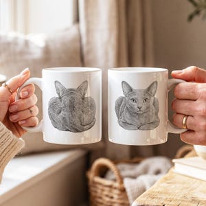 May include: Two white ceramic mugs featuring a detailed gray sketch of a cat. The left mug displays the cat from behind, showing its ears and back. The right mug shows the same cat from the front, with its face and paws visible in a resting position.