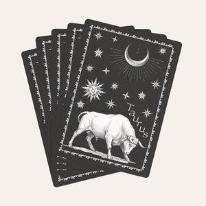 Könnte beinhalten: Ein Fächer aus rechteckigen Karten mit schwarzem Hintergrund und weißem Rand. Die oberste Karte zeigt eine weiße Illustration eines Stiers, einer Mondsichel und Sternen, mit dem Wort Taurus in weißer Schreibschrift auf der rechten Seite.