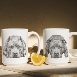 May include: Two white ceramic mugs featuring a detailed black and white pencil-style illustration of a pit bull dog lying down. The mugs have a standard cylindrical shape with a side handle. A single slice of fresh yellow lemon sits between them.