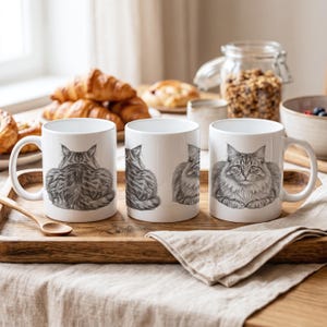 May include: Three white ceramic mugs featuring a detailed black and white sketch of a fluffy cat. The first mug shows the cat from behind, the second shows a side profile, and the third displays the cat facing forward with its paws tucked in.