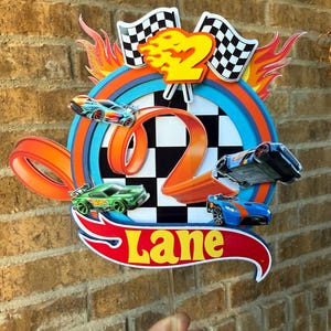 Może przedstawiać: Kolorowa dekoracja urodzinowa z motywem samochodów wyścigowych i cyfrą 2. Projekt zawiera flagę w szachownicę, płomienie i słowo "Lane" w kolorze żółtym na czerwonym banerze. Samochody są niebieskie, zielone i pomarańczowe.