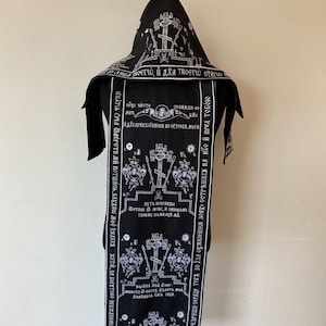 Może przedstawiać: Czarna szata religijna z białym haftowanym tekstem i symbolami religijnymi. Szata posiada kaptur i długi, prostokątny panel przedni. Tekst wydaje się być w języku słowiańskim. Projekt zawiera krzyże, anioły i inne elementy ikonografii religijnej.