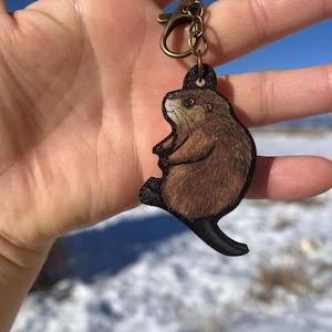 May include: A wooden keychain featuring a detailed illustration of a brown otter with a black tail. The keychain is attached to a bronze-colored clasp and chain. The text "No AI used in my Art" is visible in the upper left corner.