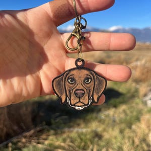 May include: A brown and black dog head keychain with a bronze-colored clasp and ring. The keychain has the text "No AI used in my Art" in the upper right corner. The dog's head is a detailed illustration with a white collar.