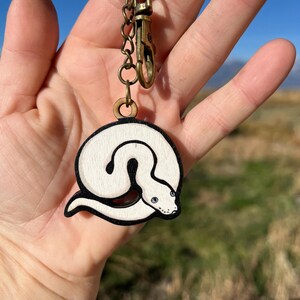 May include: A wooden keychain featuring a white snake design with black outlines and blue eyes. The keychain has a gold-colored clasp and chain. The text "No AI used in my Art" is visible in the upper right corner.