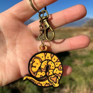 May include: A wooden keychain featuring a yellow and black snake design. The snake is coiled in a circular shape. The keychain has a bronze-colored clasp and ring. The text "No AI used in my Art" is visible in the background.