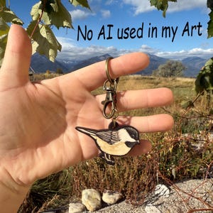 May include: A wooden bird keychain with a black and white design, attached to a bronze-colored clasp and ring. The text "No AI used in my Art" is visible in the background. The keychain is held in front of a scenic outdoor backdrop.