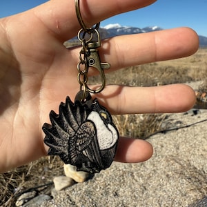 May include: A wooden keychain featuring a detailed illustration of a bird with black, white, and brown feathers. The keychain has a bronze-colored clasp and ring. The text "No AI used in my Art" is visible in the background.