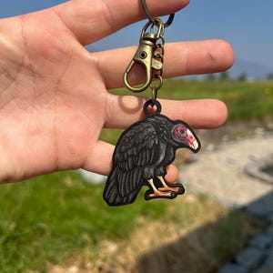May include: A black vulture keychain with a red head and yellow feet. The keychain is attached to a bronze-colored clasp and ring. The text "No AI used in my Art" is visible in the upper left corner.