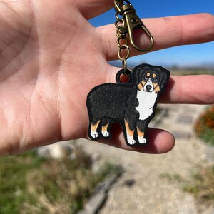 May include: A wooden keychain featuring a painted Australian Shepherd dog. The dog is black, white, and tan, with a detailed design. The keychain has a bronze-colored clasp and chain. The text "No AI used in my Art" is visible.