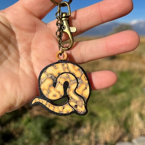 May include: A wooden keychain featuring a detailed illustration of a yellow and black snake. The snake design is circular, with a black border and a bronze-colored clasp and key ring. The text "No AI used in my Art" is visible.