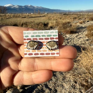 May include: A pair of frog-themed stud earrings on a patterned card. The earrings feature a detailed frog design with brown and tan tones. The card has a colorful pattern of green, orange, and white. The text "No AI used in my Art" is visible in the background.