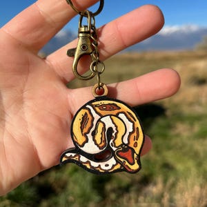 May include: A wooden keychain featuring a detailed illustration of a snake in shades of brown, yellow, and white. The keychain has a gold-colored clasp and ring. The text "No AI used in my Art" is visible in the background.