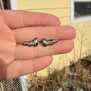 May include: A pair of small, detailed earrings featuring a reptile design with black, tan, and white accents. The earrings are held in a hand, with the text "No AI used in my Art" visible in the background.