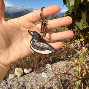 May include: A wooden keychain featuring a detailed illustration of a loon bird. The bird has a black and white patterned body, a red eye, and a yellow beak. The keychain is held up in front of a scenic mountain backdrop. The text "No AI used in my Art" is visible.