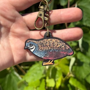 May include: A wooden keychain featuring a quail design. The bird has blue, brown, and red coloring with a white stripe on its face. The keychain has a bronze-colored clasp and key ring.