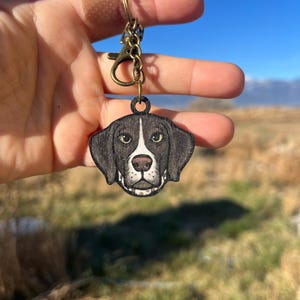May include: A wooden keychain featuring a dog's face with black and white markings, a pink nose, and green eyes. The keychain is attached to a bronze-colored clasp and chain. The text "No AI used in my Art" is visible in the background.
