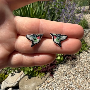 May include: A pair of stud earrings shaped like hummingbirds. Each earring features a detailed illustration of a hummingbird with a red throat, green chest, and black and white wings. The birds are depicted in a flying position with wings spread.