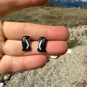 May include: A pair of black and white penguin stud earrings. The earrings are held in a hand, with a natural outdoor background. The text "No AI used in my Art" is visible in the upper right corner.