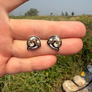 May include: A pair of stud earrings featuring a coiled snake design with a white background and brown and black patterned scales. The earrings have a smooth, glossy finish and are shaped like a rounded triangle with a pointed bottom edge.