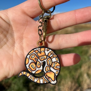 May include: A round keychain featuring a detailed illustration of a snake in shades of orange, white, and black. The keychain has a bronze-colored metal clasp and ring. The text "No AI used in my Art" is visible.