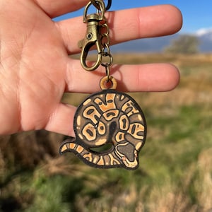 May include: A wooden keychain featuring a detailed illustration of a ball python. The snake is depicted in shades of brown, tan, and black, with a coiled body and a detailed head. The keychain includes a bronze-colored clasp and ring, and the text "No AI used in my Art".