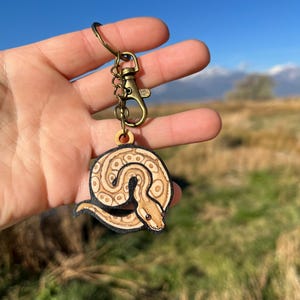 May include: A wooden keychain featuring a detailed snake design. The snake is tan and brown with a black outline, attached to a bronze-colored clasp and keyring. The text "No AI used in my Art" is visible in the background.