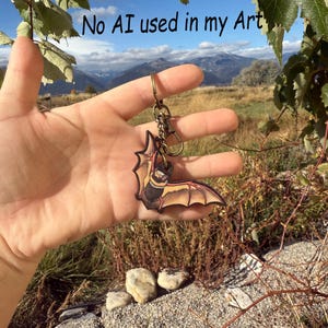 May include: A bat-shaped keychain with a bronze-colored clasp. The bat has a black body, tan wings, and red accents. The background features a scenic view of mountains and a blue sky. The text "No AI used in my Art" is visible.