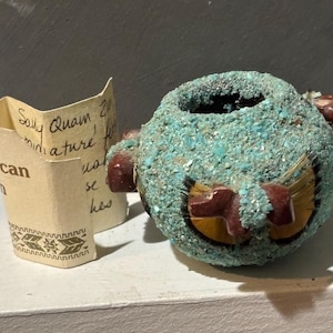 May include: A small, turquoise and brown ceramic pot with a hand-painted design. The pot is decorated with turquoise stones and has a small opening at the top. The text "Sandy Quan 20" is written on a piece of paper next to the pot.