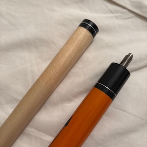 May include: A pool cue with a light brown shaft and an orange and black handle. The handle has a silver accent ring. The cue is designed for playing billiards and is shown against a white background.