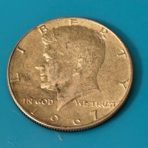 May include: A close-up of a gold-coloured Kennedy half dollar coin. The coin features a profile of John F. Kennedy and the inscriptions "IN GOD WE TRUST" and "LIBERTY". The year "1971" is visible.
