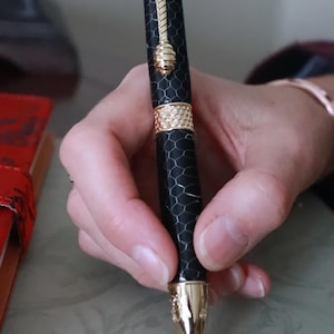 Peut inclure: Un stylo noir et doré avec un motif en nid d'abeille et des accents d'abeille. Le stylo présente une pointe dorée, une bande décorative et un motif sur le thème des abeilles sur le dessus. Le stylo est tenu dans une main.