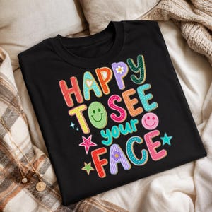 May include: A black t-shirt featuring the colorful text "HAPPY TO SEE YOUR FACE" in a playful, multi-patterned font. The design includes graphic elements such as a green smiley face, a pink smiley face, small flowers, and stars in various colors.